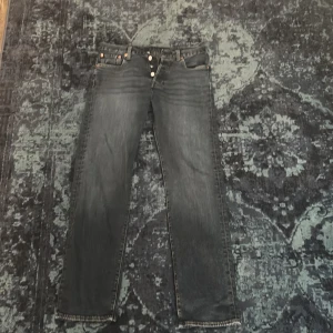 Levi's 501 svarta jeans W30 L32 - Klassiska svarta Levi's 501 jeans med raka ben och knappgylf. Jeansen har en snygg tvättad look och är tillverkade i slitstarkt bomullsjeans. Perfekta för dig som gillar tidlös stil och vill ha ett par jeans som funkar till allt.