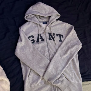 GANT vintage Zip Hoodie  - Snygg grå hoodie från GANT med stor svart logga över bröstet. Tröjan har huva med snörning, dragkedja framtill och två fickor. Mjuk insida och ribbade muddar vid ärmslut och nederkant. Perfekt för chill dagar eller när du vill ha en clean och grish look