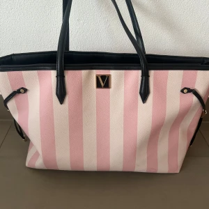 Randig shoppingväska från Victoria's Secret - Stor och rymlig shoppingväska från Victoria's Secret med rosa och vita breda ränder. Väskan har svarta handtag och detaljer, samt ett guldfärgat V-emblem framtill. Insidan är fodrad med svart tyg och har diskreta ränder. Perfekt för dig som gillar en färgglad och trendig look.