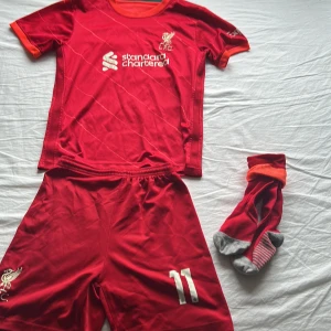 Liverpool M. Salah fotbollströja röd - Säljer en röd Liverpool fotbollströja med M. Salah och nummer 11 på ryggen. Tröjan har korta ärmar, vita detaljer och klubbmärke på bröstet. Ingår matchande shorts och röda/grå strumpor. Materialet är lätt polyester, perfekt för träning eller match.