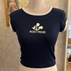 Mörkblå ribbad Mickey Mouse baby tee - Mörkblå ribbad topp från H&M x Disney med vit text och Mickey Mouse-tryck framtill. Topp med korta ärmar och kontrasterande vita kanter vid hals och ärmslut. Köpt från barnavdelningen i storlek 146/152 men passar storlek XS/S om man gillar tighta baby-tees. 