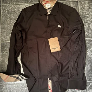 Svart Burberry skjorta med rutig detalj - Svart skjorta från Burberry i storlek S med klassiska rutiga detaljer på insidan av kragen och manschetterna. Skjortan har knappar framtill och en liten Burberry-logga broderad på bröstet. Snygg och stilren design som sticker ut med de rutiga inslagen. Priset kan diskuteras, har i Str M och I vit dessutom 