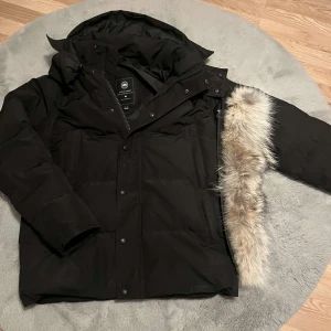 Svart dunjacka från Canada Goose - Säljer en svart dunjacka från Canada Goose med huva och avtagbar pälskant. Jackan har dragkedja och knappar framtill samt flera praktiska fickor. Perfekt för kalla vinterdagar och riktigt snygg streetstil.