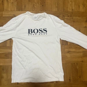 Vit långärmad tröja från Hugo Boss - Snygg vit långärmad tröja från Hugo Boss med stor svart logga framtill. Tillverkad i mjuk bomull som känns skön mot huden. Perfekt för dig som gillar stilrena och klassiska plagg med en modern touch.