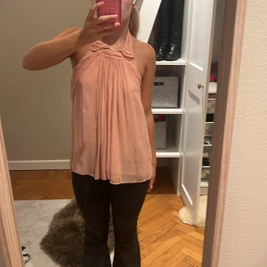 Ljusrosa omlott halterneck blus från Zara - Säljer en ljusrosa blus med halterneck och omlottdetalj vid halsen. Toppen är ärmlös och har ett flowy, löst fall med snygga veck framtill. Perfekt för dig som gillar en feminin och chill vibe. Materialet känns lätt och luftigt.