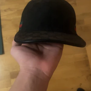 Gucci keps svart - Säljer en svart keps från Gucci med klassiska gröna och röda ränder på sidorna. Kepsen har böjd skärm och justerbart spänne bak i skinn. Materialet är troligtvis bomull med detaljer i skinn. Perfekt för dig som vill ha en stilren och lyxig accessoar.