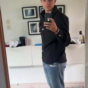 Svart overshirt från Urban Classics - Säljer en svart overshirt från Urban Classics i storlek S. Skjortan har två bröstfickor med knappar och klassisk krage. Perfekt att bära som lager över en t-shirt. Stängs med silvriga knappar framtill.