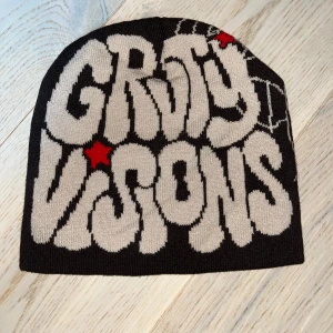 Grvty Visions brun mössa - Cool svart mössa med stor vit text 'GRITTY VISIONS' och röda stjärnor på framsidan. Baksidan har ett stort vitt spindelnätsmönster. Stickad i mjukt material, perfekt för dig som vill sticka ut med din stil.