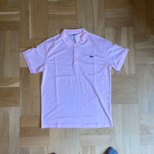 Ljusrosa Lacoste piké  - Fräsch ljusrosa piké från Lacoste med klassisk krage och korta ärmar. T-shirten har den ikoniska krokodilloggan broderad på bröstet och är gjord i mjuk bomull. Perfekt för dig som gillar stilren och sportig vibe.