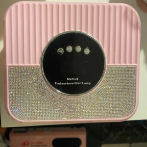  uv/nagellampa  - Superfin rosa nagellampa med glittrande strassdetaljer och svart display. Lampan har fyra knappar för olika timerinställningar och en snygg, modern design. Perfekt för att fixa gelenaglar hemma med extra bling.