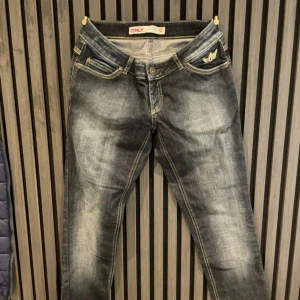 Mörkblå jeans från ONLY med brodyr - Snygga mörkblå jeans från ONLY med slitningar och kontrastsömmar. Fickan framtill har en broderad detalj med vingar och krona. Klassisk femficksmodell i stretchig denim som sitter skönt och passar till allt.
