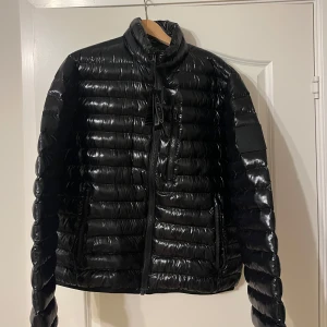 Svart dunjacka Peak Performance XL - Snygg svart dunjacka från Peak Performance i glansigt material. Jackan har quiltad design, hög krage och dragkedjor framtill samt på fickorna. Perfekt för kalla dagar och har en stilren logga på ärmen.