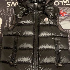 Svart dunväst från Moncler med huva - Snygg svart dunväst från Moncler med glansig finish och avtagbar huva. Västen har två fickor med dragkedja framtill och klassisk Moncler-logga på bröstet. Perfekt för lager-på-lager och riktigt street. Stängs med dragkedja hela vägen. Äkta och pris kan diskuteras!