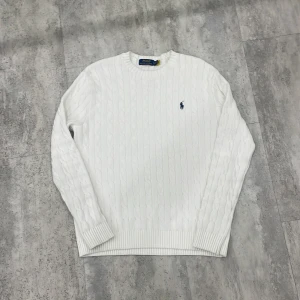Vit kabelstickad tröja Polo Ralph Lauren - Vit kabelstickad tröja från Polo Ralph Lauren med klassisk rund hals och blå broderad logga på bröstet. Tröjan har ribbade muddar och är långärmad, perfekt för lager-på-lager och snygg till jeans eller kjol.