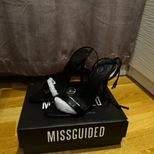 Svarta klackar från Missguided, strl 36 - Snygga svarta klackar från Missguided i storlek 36. Skorna har spetsig tå, hög klack och en tunn rem över tårna samt runt vristen. Tillverkade i glansigt syntetmaterial som ger en stilren look. Perfekta för att lyfta din outfit. Helt nya och säljer på grund av stor i storlek:) 