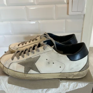 Golden Goose Superstar sneakers vit/grå - Golden Goose Superstar sneakers i vitt skinn med grå mockadetaljer och stjärna på sidan. Grå snörning, blått hälparti och beige sula med vintage-look. Klassisk låg modell med rund tå och platt sula, perfekt för en avslappnad streetstyle. Box och allt og medkommer