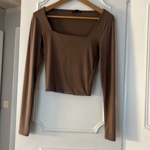 Brun långärmad topp med fyrkantig ringning - Snygg brun långärmad topp med croppad passform och fyrkantig ringning. Tillverkad i ett stretchigt, mjukt material som sitter tight mot kroppen. Perfekt att styla med jeans eller kjol för en trendig look.