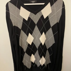 Svart och grå rutig tröja Madison Avenue - Snygg svart tröja från Madison Avenue med klassiskt rutig argyle-mönster i grått och vitt. V-ringad hals och långa ärmar. Perfekt för dig som gillar stilrena och tidlösa plagg med lite preppy vibe. Materialet är mjukt och stickat för extra komfort.