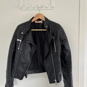 Svart skinnjacka  - Snygg svart skinnjacka från Chiquelle med coola detaljer som quiltade axlar, dragkedjor och bälten i sidorna. Jackan har långärmade ärmar och en klassisk bikerstil med asymmetrisk dragkedja. Perfekt för dig som vill ha en edgy look.