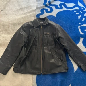 Svart skinnjacka från Saki - Svart skinnjacka från SAKI. Äkta skinn i bra skick. Liten repa på ryggen, se sista bilden.  Nypris: 1000 kr  Chest: 134 cm Shoulder width: 54 cm Sleeve length: 65 cm Back length: 73 cm