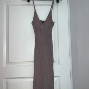 Beige ribbad långklänning med v-ringning - Snygg beige långklänning med smala axelband och djup v-ringning. Klänningen är ribbad och har en tight passform som framhäver figuren. Perfekt för dig som gillar minimalistisk och stilren design.