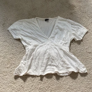 Vit omlottopp - Supersöt vit omlottopp från Gina Tricot i storlek XS. Toppen har korta ärmar, v-ringning och knytband i midjan som ger en snygg siluett. Tillverkad i ett lätt och luftigt bomullstyg, perfekt för alla väder.