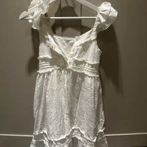 Vit klänning från Zara - Supersöt vit kortklänning från Zara med broderade blommor, volangdetaljer och knappar framtill. Klänningen har ärmlös design med romantiska axelvolanger och är gjord i mjuk bomull, perfekt för varma dagar. Passar dig som gillar en feminin och chill stil.