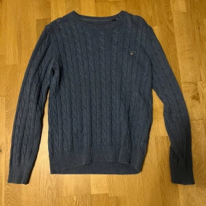 Blå kabelstickad tröja Gant - Kabelstickad blå tröja från Gant med rund halsringning och diskret logga på bröstet. Tröjan har långa ärmar och ribbade muddar vid ärmslut och nederkant. Perfekt för dig som gillar klassisk och stilren look. Size small