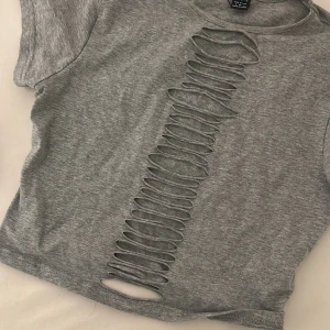 Grå t-shirt med slitsad front - Grå t-shirt från Bershka med korta ärmar och coola slitsade detaljer längs hela framsidan. T-shirten har en rund hals och är gjord i mjuk bomull som sitter skönt. Perfekt för dig som gillar edgy och unik stil.