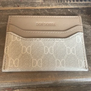 Beige kortfodral från Don Donna - Snyggt och stilrent kortfodral från Don Donna i beige med diskret mönster av DD-loggor. Fodralet har flera kortfack och är gjort i ett syntetmaterial med läderlook. Perfekt för dig som vill ha koll på korten utan att ta plats i fickan.