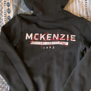 Svart hoodie från McKenzie - Svart hoodie från McKenzie med tryck i vitt och rött framtill. Klassisk huva med snörning och känguruficka. Mjuk insida och långärmad modell, perfekt för en chill och sportig stil. Mycket bra skick. Pris kan diskuteras vid snabb affär