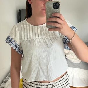Vit blus med blå broderi - Supersöt vit blus med korta ärmar och blå broderade detaljer längs ärmsluten. Blusen har en lös passform, rund halsringning och fina veck upptill. Perfekt för en fräsch och somrig look.