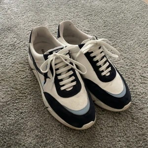 Axel Arigato sneakers - Snygga sneakers från Axel Arigato i vitt och marinblått med detaljer i ljusblått. Skorna har snörning, rund tå och en cool fågelbroderi på sidan. Tillverkade i skinn och mocka för en lyxig känsla. Perfekta för dig som gillar stilrena och trendiga sneakers.