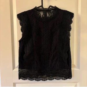 Svart spetsblus från ONLY - Supersnygg svart blus från ONLY med spetsdetaljer och vackert mönstrad mesh. Ärmlös modell med volangkant längs axlar och nederkant. Perfekt för dig som gillar romantiska och detaljerade plagg. 