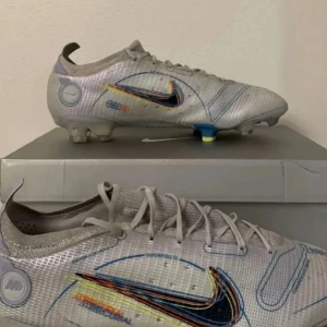 Nike Mercurial fotbollsskor silver - Snygga Nike Mercurial fotbollsskor i silver med färgglada detaljer och svart swoosh. Skorna har snörning, platt sula med dobbar och är tillverkade i syntetmaterial. Perfekta för dig som vill sticka ut på planen.