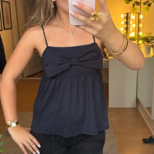 Mörkblå linnetopp med rosett - Supersöt mörkblå linnetopp med smala axelband och stor rosett framtill. Toppen har en lätt rynkad, luftig passform och är perfekt för varma dagar. Materialet känns mjukt och svalt mot huden. Enkel att styla med både jeans och kjol. Sitter som en xs/s. Nästintill aldrig använd i väldigt fint skick. Läs bio innan köp.