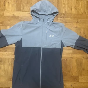 Grå vindjacka från Under Armour - Snygg tvåfärgad vindjacka från Under Armour i ljusgrått och mörkgrått. Jackan har huva, hel dragkedja och två sidofickor. Perfekt för blåsiga dagar och sportiga outfits. Under Armour-logga på bröstet ger en sportig vibe.