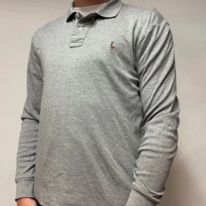 Grå långärmad pikétröja Polo Ralph Lauren - Snygg grå långärmad pikétröja från Polo Ralph Lauren med klassisk krage, knappar och broderad logga på bröstet. Tillverkad i mjuk bomull med Pima Soft Touch för extra komfort. Perfekt för en clean och stilren look.