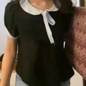 Svart blus med vit krage - Supersöt svart blus med kontrasterande vit krage och knytrosett framtill. Blusen har korta ärmar med vita kanter och en lös passform som ger en chill och trendig vibe. Perfekt för dig som gillar en söt och stilren look.