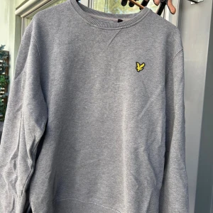 Grå sweatshirt från Lyle & Scott - Klassisk grå sweatshirt från Lyle & Scott med rund hals och broderad gul logga på bröstet. Tröjan har ribbade muddar vid ärmslut och nederkant, och är tillverkad i mjuk bomull. Perfekt för en chill och stilren look.