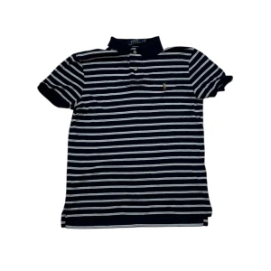 Randig Polo Ralph Lauren t-shirt - Snygg marinblå och vit randig t-shirt från Polo Ralph Lauren med klassisk polokrage och broderad logga på bröstet. Modellen är custom slim fit med korta ärmar och knappar vid halsen. Perfekt för dig som gillar stilren och sportig look.