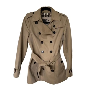 Beige trenchcoat från Burberry - Säljer en klassisk beige trenchcoat från Burberry, modell The Kensington. Jackan har dubbelknäppning med mörka knappar och ett bälte i midjan. Den har en ikonisk rutig insida och är perfekt för höst och vår. Storlek 34