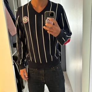 Randig stickad tröja från McNeal - Snygg marinblå stickad tröja från McNeal med vita vertikala ränder och röda armbågslappar. V-ringad modell med lång ärm och broderad logga på bröstet. Perfekt för dig som gillar klassisk men ändå sportig stil.