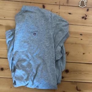 Grå t-shirt från GANT i bomull - Snygg grå t-shirt från GANT med klassisk rund hals och broderad logga på bröstet. T-shirten är enkel och stilren, perfekt till jeans eller shorts. Tillverkad i mjuk bomull för skön känsla hela dagen.