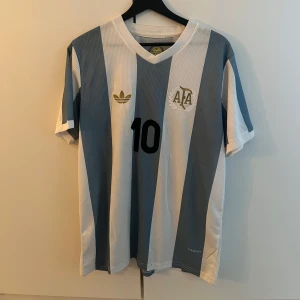Argentina Messi fotbollströja Adidas - Säljer en klassisk Argentina fotbollströja från Adidas med Messi och nummer 10 på ryggen. Tröjan har breda ljusblå och vita ränder, korta ärmar och v-ringad krage. Materialet är lätt och ventilerande polyester, perfekt för match eller träning. Replika
