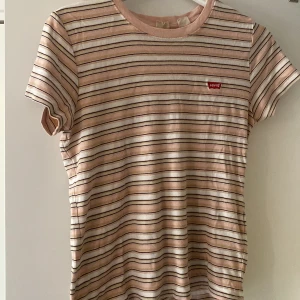 Randig t-shirt från Levi's i beige - Snygg t-shirt från Levi's med tunna horisontella ränder i beige, vitt och svart. Klassisk rund hals och korta ärmar. Mjuk bomullskvalitet och liten röd Levi's-logga på bröstet. Perfekt till jeans eller shorts för en chill look.