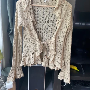Beige virkad blus från Kookai - Superfin beige virkad blus från Kookai med långa ärmar, volangkant och knyt framtill. Blusen har virkade detaljer och små träpärlor längs krage och volanger. Perfekt för dig som gillar boho-stil och unika plagg.