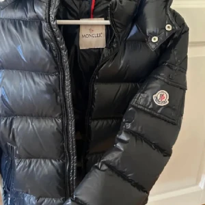 Svart Moncler dunjacka med huva - Snygg svart dunjacka från Moncler med glansig finish och klassisk logga på ärmen. Jackan har huva, dragkedja och quiltad design som håller värmen. Perfekt för kalla dagar och riktigt stilren att bära till det mesta.