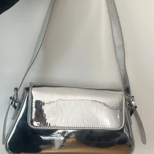 Silvrig axelväska i syntet - Snygg axelväska i metallic-silver med spegelblank finish och stilren design. Väskan har ett stort fack med dragkedja och innerfickor för småprylar. Perfekt för dig som vill sticka ut med en futuristisk look. Justerbar axelrem för skön passform.