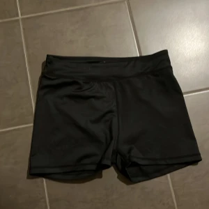 Svarta träningsshorts från Pro Touch - Svarta, tighta träningsshorts från Pro Touch i storlek 164. De är tillverkade i ett stretchigt syntetmaterial och har snörning i midjan för bästa passform. Perfekta för dig som vill ha bekväma och stilrena shorts till gymmet eller löprundan.